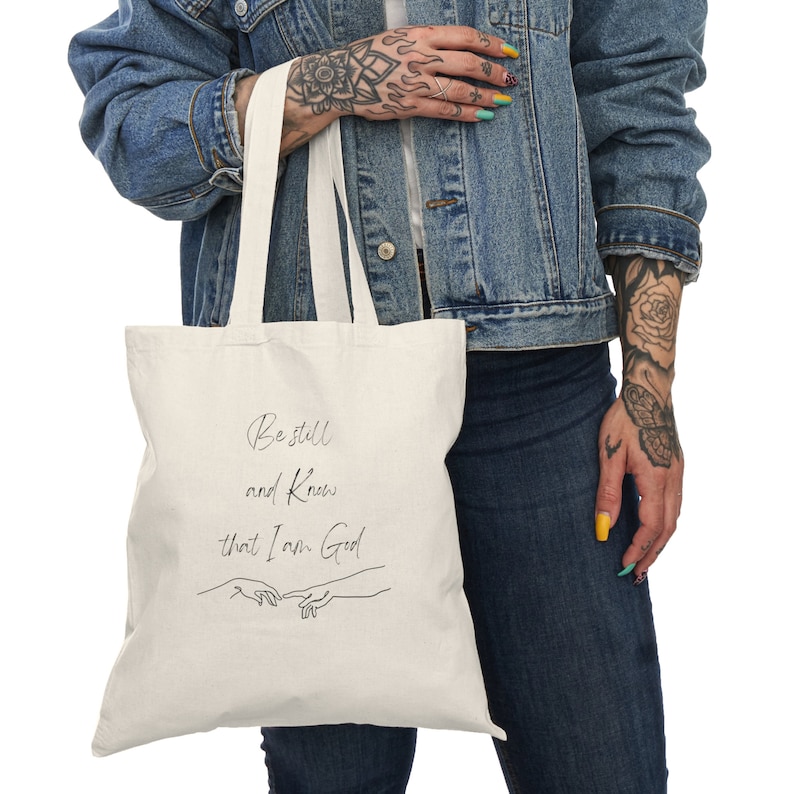 May include: A white canvas tote bag with black text that reads "Be still and know that I am God". The text is in a cursive font and is accompanied by a simple line drawing of two hands reaching towards each other.