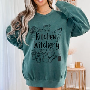 Może przedstawiać: Zielony sweter w kolorze turkusowym z napisem "Kitchen Witchery" czarnymi literami. Projekt zawiera r&oacute;żne ilustracje związane z kuchnią, takie jak moździerz i tłuczek, wałek do ciasta, n&oacute;ż i trzepaczka.