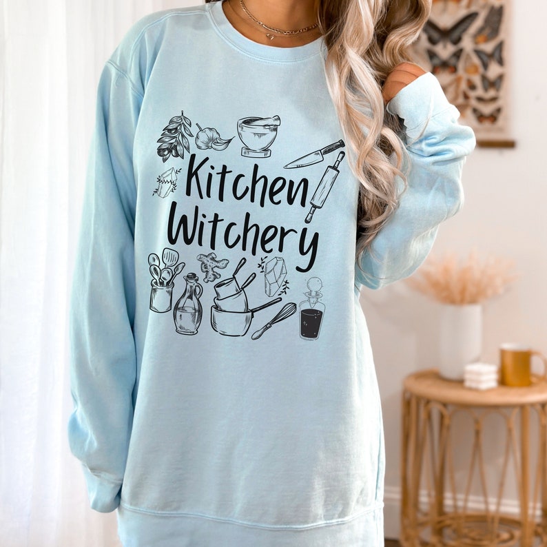 Może przedstawiać: Jasno błękitna bluza z napisem "Kitchen Witchery" w czarnych literach. Bluza zawiera wz&oacute;r r&oacute;żnych narzędzi kuchennych i składnik&oacute;w, w tym wałek do ciasta, trzepaczkę, butelkę, miskę i kryształ.