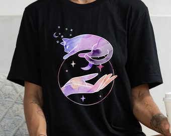 Op de afbeelding: Een zwart T-shirt met een aquarelontwerp van een paarse kat die boven een hand zweeft met een halve maan en sterren op de achtergrond.