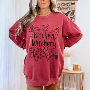 Może przedstawiać: Czerwona bluza z czarnym nadrukiem graficznym z napisem "Kitchen Witchery" i przedstawiającym kuchenne narzędzia i zioła.