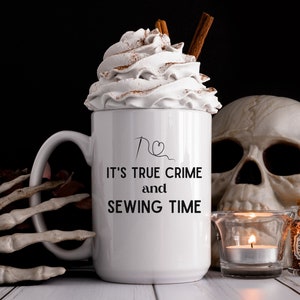 May include: White ceramic mug with a black design that says "It's True Crime and Sewing Time" with a needle and thread heart graphic. The mug is filled with whipped cream and a cinnamon stick. The mug is sitting on a white surface with a skull and a candle.