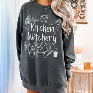 Może przedstawiać: Szara bluza z kapturem z napisem "Kitchen Witchery" w kolorze białym. Bluza zawiera r&oacute;żne ilustracje związane z kuchnią, w tym moździerz i tłuczek, wałek do ciasta, n&oacute;ż, trzepaczkę, butelkę i kryształ.