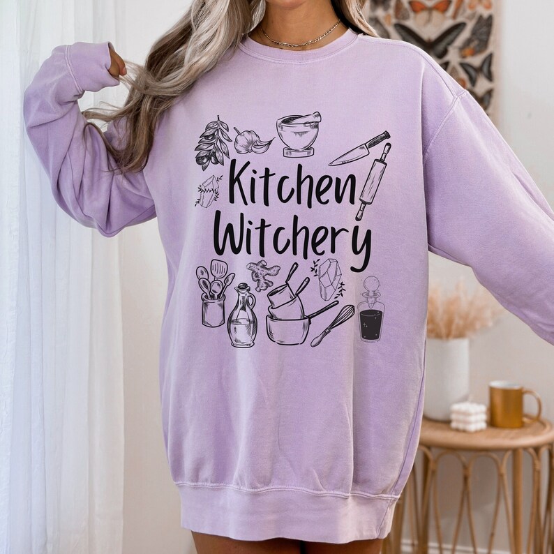 Może przedstawiać: Lavenדרowy sweter z nadrukiem "Kitchen Witchery" na przodzie. Tekst jest otoczony r&oacute;żnymi ilustracjami związanymi z kuchnią, w tym moździerzem i tłuczkiem, wałkiem do ciasta, nożem, trzepaczką i butelką oleju.