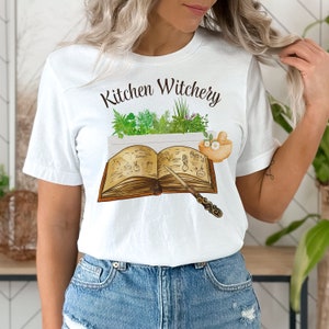 May include: White t-shirt with the text "Kitchen Witchery" printed on the front. The design features a book with a wand, herbs, and a mortar and pestle.