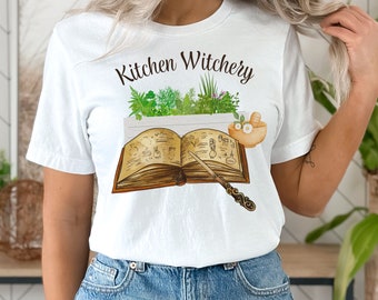 May include: White t-shirt with the text "Kitchen Witchery" printed on the front. The design features a book with a wand, herbs, and a mortar and pestle.