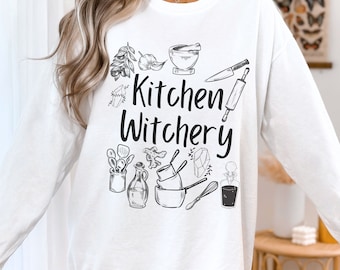 May include: White long-sleeve sweatshirt with a black graphic design that says "Kitchen Witchery" and features kitchen utensils, herbs, and crystals.