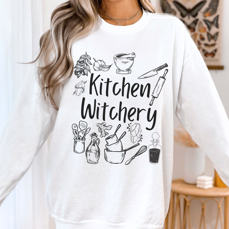 Może przedstawiać: Biała bluza z długim rękawem z czarnym nadrukiem graficznym, na kt&oacute;rym widnieje napis "Kitchen Witchery" oraz przedstawione są kuchenne narzędzia, zioła i kryształy.