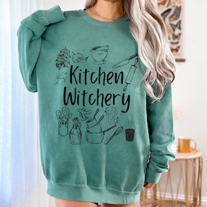 Może przedstawiać: Zielony sweter w kolorze turkusowym z nadrukiem "Kitchen Witchery" w kolorze czarnym. Wz&oacute;r zawiera r&oacute;żne przybory kuchenne i składniki, takie jak wałek do ciasta, n&oacute;ż, moździerz i tłuczek oraz zioła.