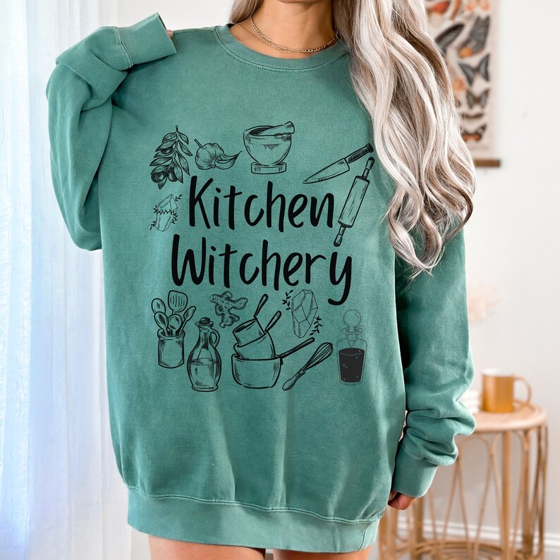 Może przedstawiać: Zielony sweter w kolorze turkusowym z nadrukiem "Kitchen Witchery" w kolorze czarnym. Wz&oacute;r zawiera r&oacute;żne przybory kuchenne i składniki, takie jak wałek do ciasta, n&oacute;ż, moździerz i tłuczek oraz zioła.