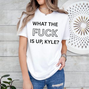 May include: White t-shirt with the text "WHAT THE FUCK IS UP, KYLE?" in black block letters. The shirt is a crew neck style and appears to be made of a soft material. The shirt is being worn with blue jeans.