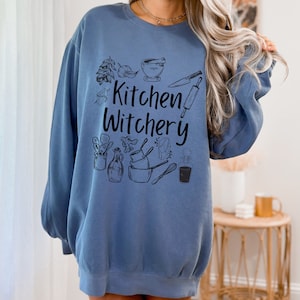 Może przedstawiać: Niebieska bluza z nadrukiem "Kitchen Witchery" czarnymi literami. Bluza zawiera r&oacute;żne ilustracje związane z kuchnią, w tym moździerz i tłuczek, wałek do ciasta, n&oacute;ż i butelkę oleju.