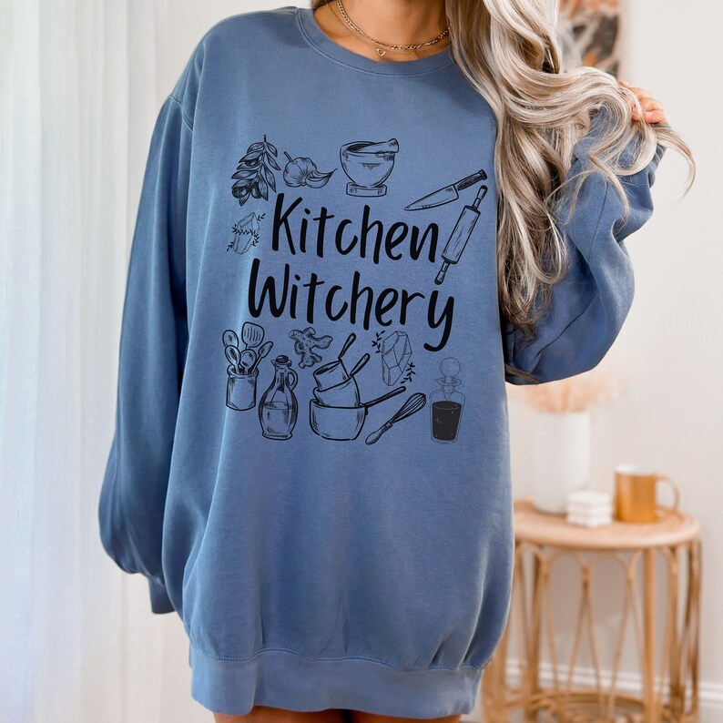 Może przedstawiać: Niebieska bluza z nadrukiem "Kitchen Witchery" czarnymi literami. Bluza zawiera r&oacute;żne ilustracje związane z kuchnią, w tym moździerz i tłuczek, wałek do ciasta, n&oacute;ż i butelkę oleju.