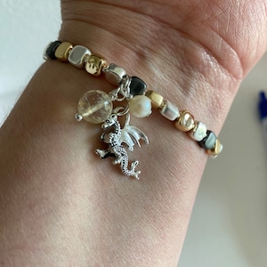 May include: A silver charm bracelet with a dragon charm, a clear quartz bead, and a white pearl bead. The bracelet is made of silver, gold, and black beads.
