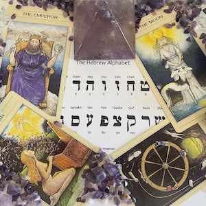 Könnte beinhalten: Tarotkarten mit Bildern von Der Stern, Der Kaiser, Der Mond und Das Rad des Schicksals. Die Karten liegen auf einer weißen Oberfläche mit verstreuten Amethystkristallen. Ein Blatt Papier mit dem hebräischen Alphabet ist in der Mitte des Bildes sichtbar.