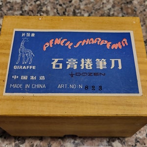 Puede incluir: Una caja azul con una ilustración de jirafa y el texto "Pencil Sharpener" en un círculo rosa. La caja también tiene el texto "石膏 鉛筆" en negro y el texto "Made in China" en blanco. La caja está hecha de madera y tiene un color marrón claro.