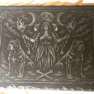 May include: A rectangular, dark gray decorative slate featuring a central image of a woman holding torches and keys, flanked by two dogs. The design includes moons, stars, and ornate borders, creating a mystical theme.