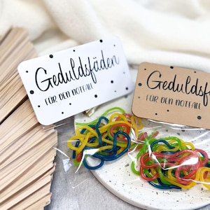 Pode incluir: Duas pequenas sacolas de presente com doces coloridos dentro. As sacolas são etiquetadas com "Geduldsfaden fur den Notfall", que se traduz como "Fio de paciência para emergências".