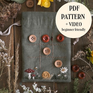 Könnte beinhalten: Ein olivgrüner Stoffbeutel mit einem gestickten Tic-Tac-Toe-Gitter und Holzknöpfen als Spielfiguren. Der untere Bereich zeigt gestickte Pilze, eine Schnecke und Gänseblümchen. Der Text auf dem Bild lautet PDF PATTERN + VIDEO beginner friendly.