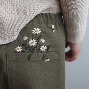 Könnte beinhalten: Olivgrüne Hose mit einer Tasche, die mit weißen Gänseblümchen und einer kleinen Biene bestickt ist. Die Hose ist aus einem strukturierten Stoff gefertigt und wird mit einem hellbeigen Pullover kombiniert.