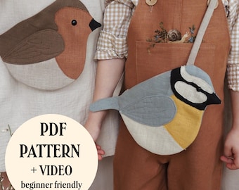 May include: A fabric bird-shaped bag featuring a blue, yellow, and black design with a long strap. A circular white graphic overlay displays the text PDF PATTERN + VIDEO beginner friendly. The bag is worn over a brown overall garment.