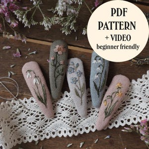 Könnte beinhalten: Fünf stoffbezogene Haarspangen mit handgestickten Blumenmotiven in Rosa, Grün, Blau und Grau. Der Text auf dem Bild lautet PDF PATTERN + VIDEO beginner friendly. Die Spangen sind auf einer weißen Spitzenborte angeordnet.