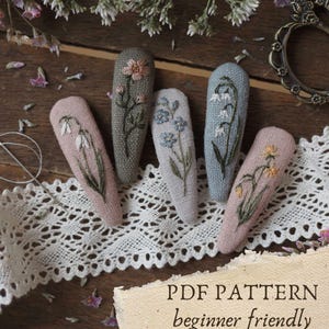 May include: Five embroidered fabric hair clips in shades of pink, gray, and blue, each featuring a different floral design. The clips are arranged on a wooden surface with lace and dried flowers. The text "PDF PATTERN beginner friendly" is visible.