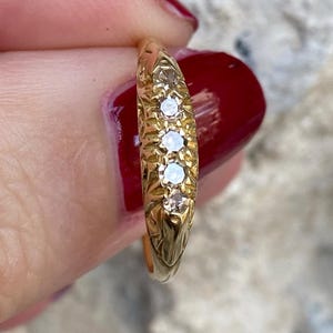 May include: A gold ring with four small, clear gemstones set in a row. The ring has an ornate, engraved design along the band. The ring is held up by a person with red nail polish.