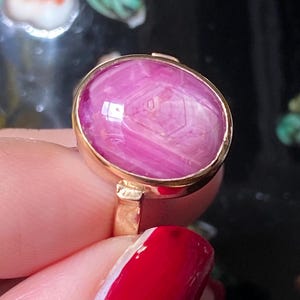 Op de afbeelding: Een gouden ring met een grote, ovale roze edelsteen. De edelsteen heeft een uniek intern patroon. De ring wordt vastgehouden door een persoon met rode nagellak. De achtergrond is donker met bloemmotieven.