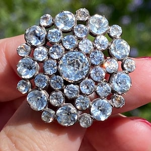May include: A round, antique brooch featuring numerous clear, sparkling gemstones. The brooch has a central, large stone surrounded by smaller stones, creating a dazzling, circular design. The gemstones are set in a silver or platinum metal.