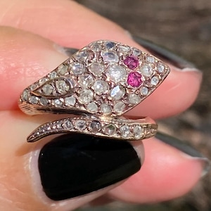 May include: A gold-toned snake ring with a head and body encrusted with small, clear gemstones. The snake's eyes are two small, bright pink gemstones. The ring is held up by a person's hand.