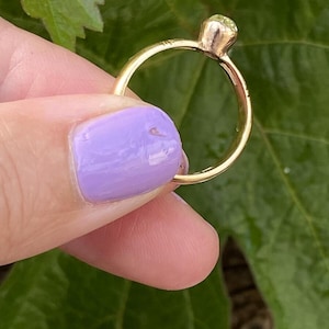 K&ouml;nnte beinhalten: Ein goldener Ring mit einem runden, hellgr&uuml;nen Edelstein. Der Ring wird vor einem Hintergrund aus gr&uuml;nen Bl&auml;ttern gehalten. Das Band des Rings ist d&uuml;nn und schlicht, und der Edelstein ist in einer Krappenfassung gefasst.