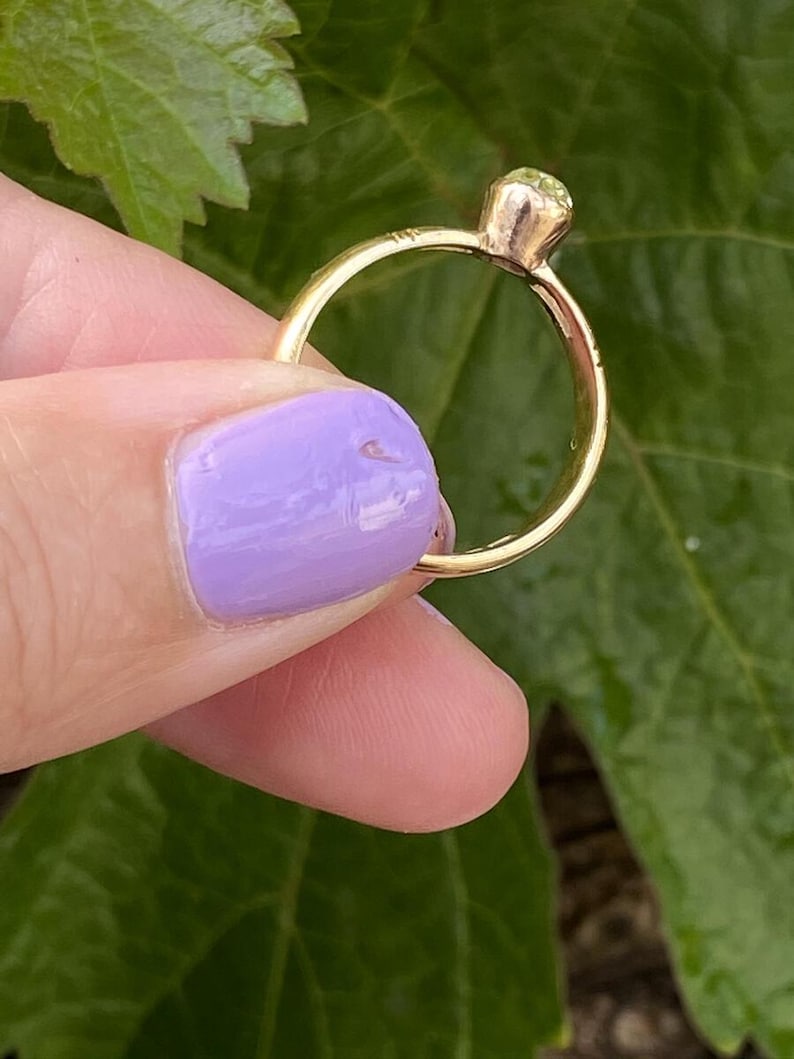 K&ouml;nnte beinhalten: Ein goldener Ring mit einem runden, hellgr&uuml;nen Edelstein. Der Ring wird vor einem Hintergrund aus gr&uuml;nen Bl&auml;ttern gehalten. Das Band des Rings ist d&uuml;nn und schlicht, und der Edelstein ist in einer Krappenfassung gefasst.