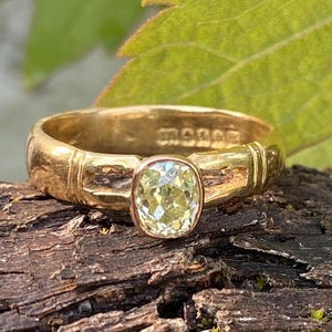 K&ouml;nnte beinhalten: Ein goldener Ring mit einem hellgelben, kissenf&ouml;rmigen Edelstein. Der Ring hat ein strukturiertes Band mit eingravierten Details und eine L&uuml;nettenfassung. Der Ring wird auf Holz mit einem gr&uuml;nen Blatt im Hintergrund pr&auml;sentiert.