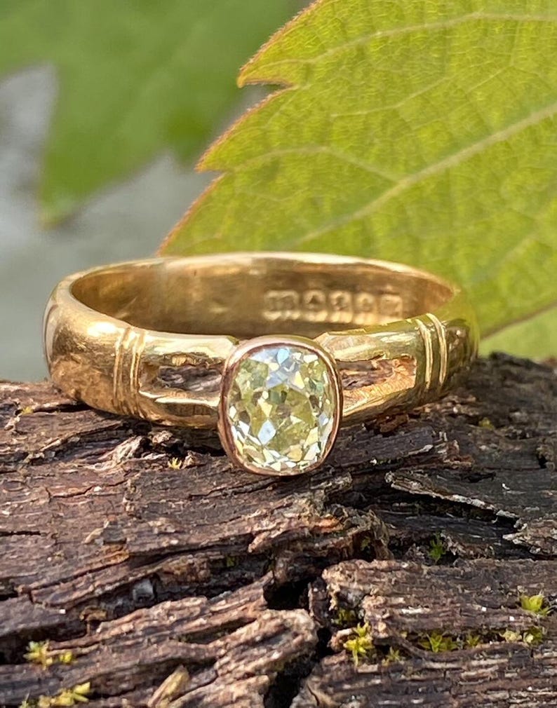 K&ouml;nnte beinhalten: Ein goldener Ring mit einem hellgelben, kissenf&ouml;rmigen Edelstein. Der Ring hat ein strukturiertes Band mit eingravierten Details und eine L&uuml;nettenfassung. Der Ring wird auf Holz mit einem gr&uuml;nen Blatt im Hintergrund pr&auml;sentiert.