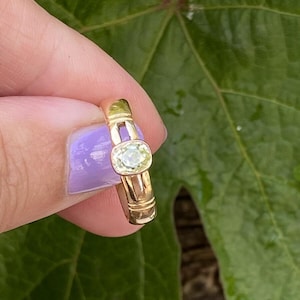 K&ouml;nnte beinhalten: Ein goldener Ring mit einem hellgr&uuml;nen, oval geformten Edelstein. Der Ring hat ein schlichtes Band mit einem rechteckigen Design an den Seiten. Der Edelstein ist in einer L&uuml;nettenfassung gefasst. Der Ring wird vor einem gr&uuml;nen Blatt gehalten.