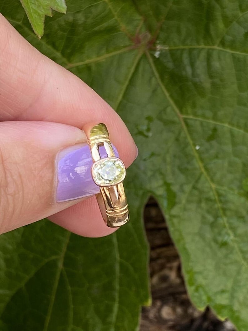 K&ouml;nnte beinhalten: Ein goldener Ring mit einem hellgr&uuml;nen, oval geformten Edelstein. Der Ring hat ein schlichtes Band mit einem rechteckigen Design an den Seiten. Der Edelstein ist in einer L&uuml;nettenfassung gefasst. Der Ring wird vor einem gr&uuml;nen Blatt gehalten.