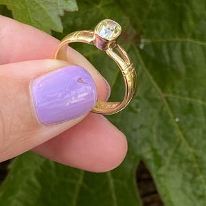 K&ouml;nnte beinhalten: Ein goldener Ring mit einem hellfarbigen Edelstein. Der Ring wird vor einem Hintergrund aus gr&uuml;nen Bl&auml;ttern gehalten. Das Band des Rings hat dekorative Details.