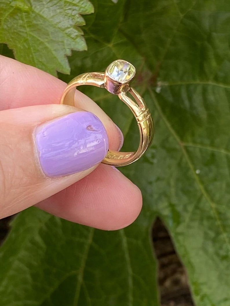 K&ouml;nnte beinhalten: Ein goldener Ring mit einem hellfarbigen Edelstein. Der Ring wird vor einem Hintergrund aus gr&uuml;nen Bl&auml;ttern gehalten. Das Band des Rings hat dekorative Details.