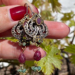May include: Ornate silver earrings with gold accents, featuring ruby and emerald gemstones. The earrings have a filigree design with dangling ruby teardrops and clusters of emerald beads. The earrings are held up against a background of green leaves.