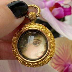 Puede incluir: Un colgante redondo dorado con un borde grabado ornamentado. El colgante presenta un retrato en miniatura del rostro de una mujer bajo una cubierta de vidrio transparente. El cierre y la fianza también son dorados. El fondo incluye flores rosas y blancas.