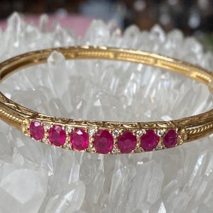 Może przedstawiać: Złota bransoletka z zawiasami z rzędem owalnych, żywych czerwonych kamieni szlachetnych i małymi diamentami. Bransoletka ma grawerowany wzór wzdłuż krawędzi.