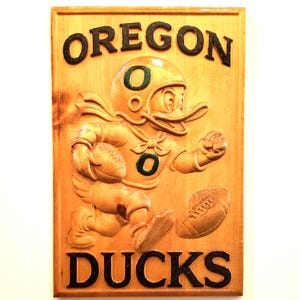 May include: Wooden plaque featuring a carved image of the Oregon Ducks mascot, a duck wearing a football helmet and uniform, running with a football. The word "OREGON" is at the top, and "DUCKS" is at the bottom, both in dark letters.