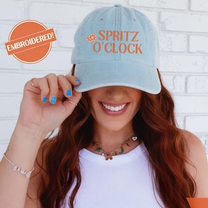 May include: Light blue baseball cap with the text "Spritz O'Clock" in orange. The cap has an embroidered design and is labeled "Embroidered!" in an orange circle. The bottom right corner of the image is labeled "Lt. Blue".