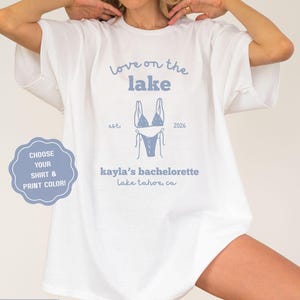 May include: White t-shirt with "Love on the Lake" and "kayla's bachelorette" text in ice blue. A bikini graphic is centered below the text. The shirt also has the text "Lake Tahoe, CA".