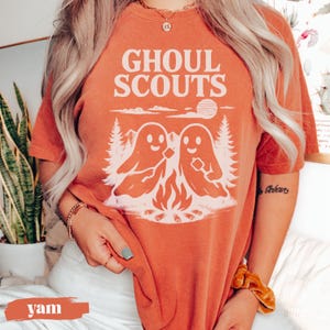 Puede incluir: Camiseta naranja con el texto "GHOUL SCOUTS" sobre un gr&aacute;fico de dos figuras de fantasmas asando malvaviscos sobre una fogata. El dise&ntilde;o incluye &aacute;rboles y un sol. La camiseta es de color naranja liso.