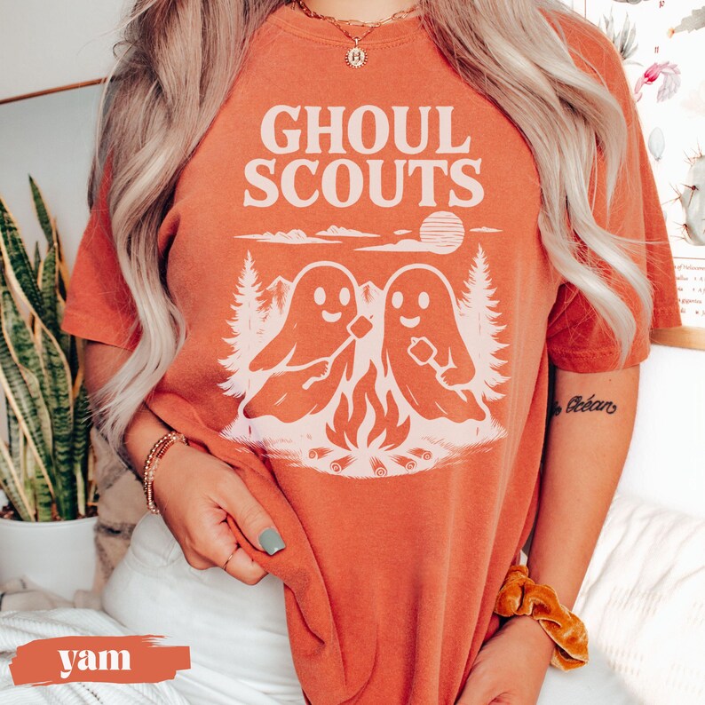 Puede incluir: Camiseta naranja con el texto "GHOUL SCOUTS" sobre un gr&aacute;fico de dos figuras de fantasmas asando malvaviscos sobre una fogata. El dise&ntilde;o incluye &aacute;rboles y un sol. La camiseta es de color naranja liso.