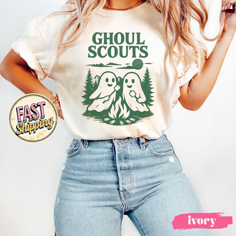 Puede incluir: Camiseta de color crema con un gr&aacute;fico verde de dos figuras de fantasmas asando malvaviscos sobre una fogata, con el texto "GHOUL SCOUTS" arriba. El dise&ntilde;o incluye &aacute;rboles y un sol. La camiseta es de estilo casual y unisex.