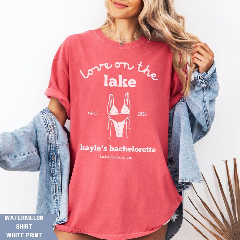 May include: Watermelon-colored shirt with white print. The shirt features the text "Love on the lake" above a bikini graphic, with "est. 2026" and "kayla's bachelorette, lake tahoe, ca" below. The shirt is a casual style.