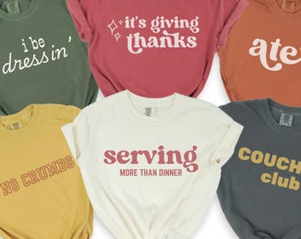 May include: A collection of six short-sleeved t-shirts in various colors, each with a different text design. The shirts are in shades of green, red, orange, yellow, white, and gray. The text includes phrases like "I be dressin'" and "It's giving thanks."
